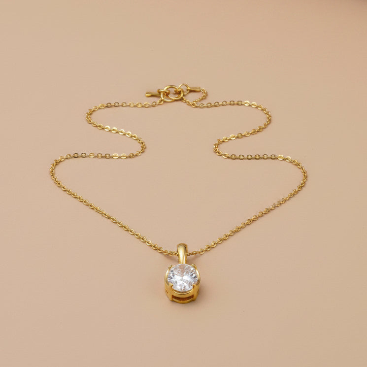 Gold necklace with a clear pendant on a brown fabric surface, with a book in the background.