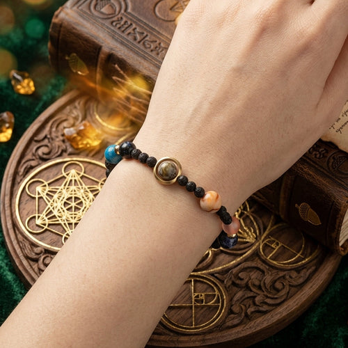 A beaded bracelet with black beads, a central oval metallic charm, and accent beads in blue and peach, worn on a person's wrist. The hand is posed over an antique-style book and occult-themed props on a dark green velvet background.