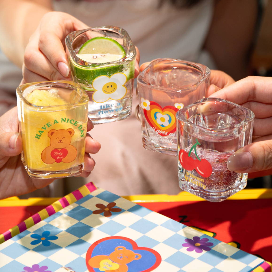 Four small clear glass shot glasses with colorful cartoon designs are being held up by hands in a toast-like arrangement. The glasses feature playful graphics including a yellow bear with the text 'HAVE A NICE DAY,' a white flower with yellow petals, a rainbow heart, and red cherries.