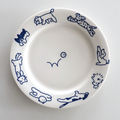 A white ceramic plate with blue line drawings of various dogs around the rim and a small blue flower motif in the center, shown from a top-down view on a light gray background.