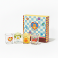 A set of four clear shot glasses with colorful cartoon bear and flower designs, arranged on a wooden cutting board next to a yellow and blue patterned gift box.
