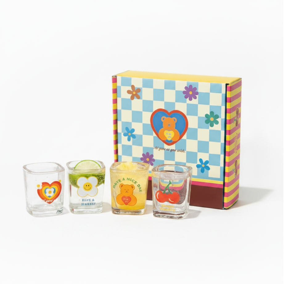 A set of four clear shot glasses with colorful cartoon bear and flower designs, arranged on a wooden cutting board next to a yellow and blue patterned gift box.