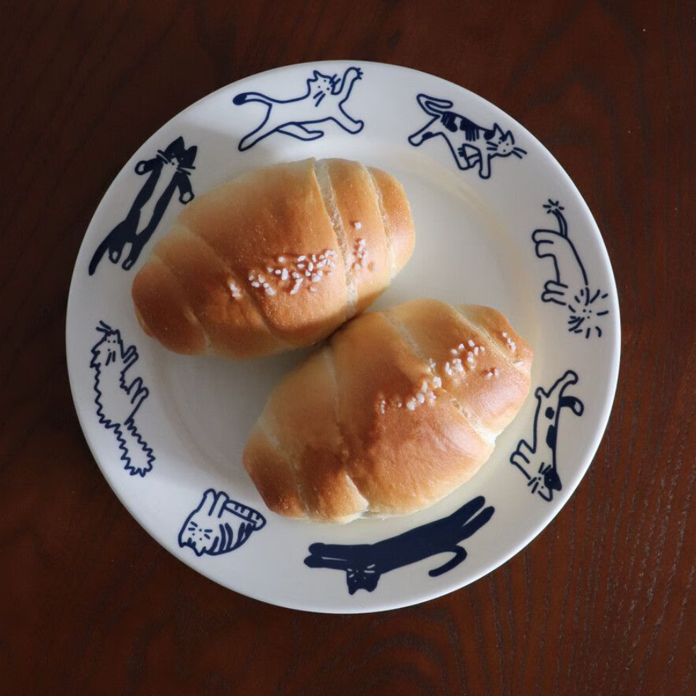 Two golden-brown baked rolls with coarse sea salt sprinkles, served on a white ceramic plate with blue cat illustrations, set on a dark wooden table.