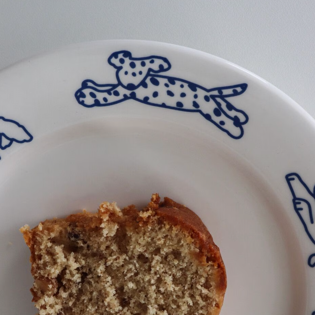 A close-up of a slice of moist banana bread on a white ceramic plate with blue line-art dog illustrations.