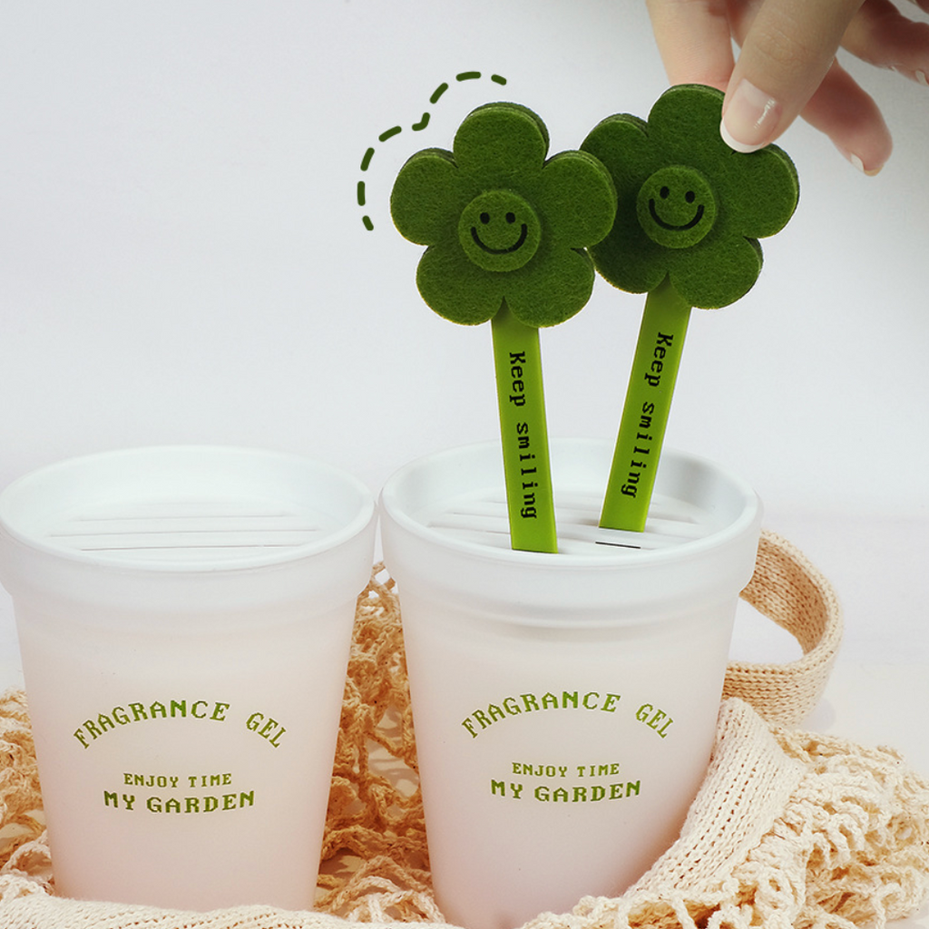 Two white plastic cups labeled 'FRAGRANCE GEL ENJOY TIME MY GARDEN' sit on a beige woven mat. Two green flower-shaped sticks with smiling faces and the words 'Keep smiling' are being inserted into the cups, with a hand holding one flower above the right cup.
