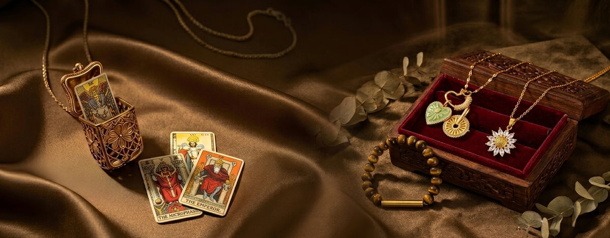 A styled flat-lay of jewelry and tarot card accessories on draped brown satin fabric. The set includes a gold locket necklace, a wooden box with a red velvet interior holding a heart-shaped pendant and a sunburst pendant, a beaded bracelet, and three tarot cards.