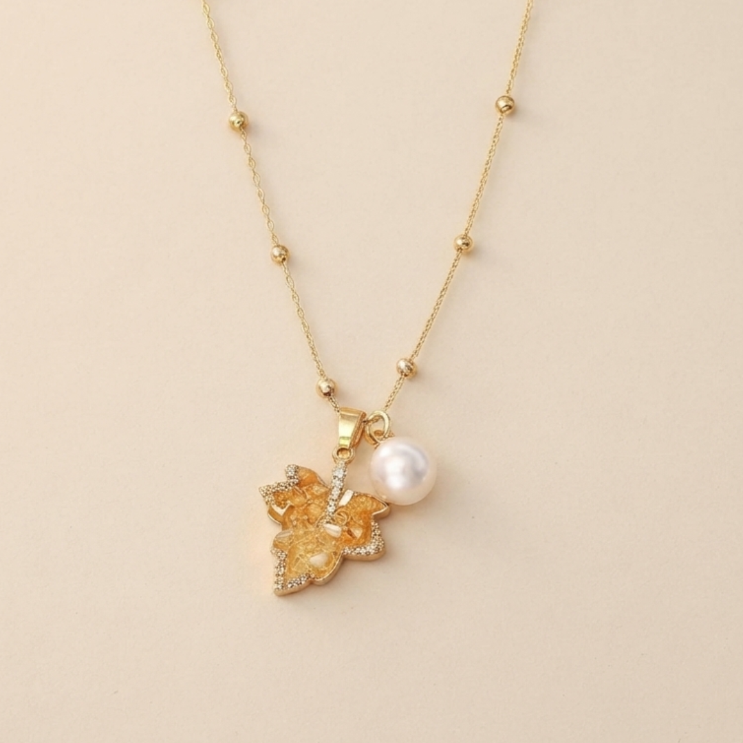 A delicate gold chain necklace featuring a small pendant with a textured yellow-orange flower, a single white pearl, and tiny clear rhinestones, displayed on a warm beige background.