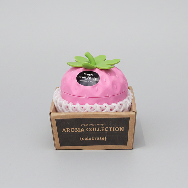 A hand holds a pink strawberry-shaped perfume box labeled 'Fresh Fruit Party' in a brown cardboard display box. The box has a white lace-like trim and reads 'AROMA COLLECTION {celebrate}'.