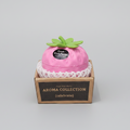 A hand holds a pink strawberry-shaped perfume box labeled 'Fresh Fruit Party' in a brown cardboard display box. The box has a white lace-like trim and reads 'AROMA COLLECTION {celebrate}'.