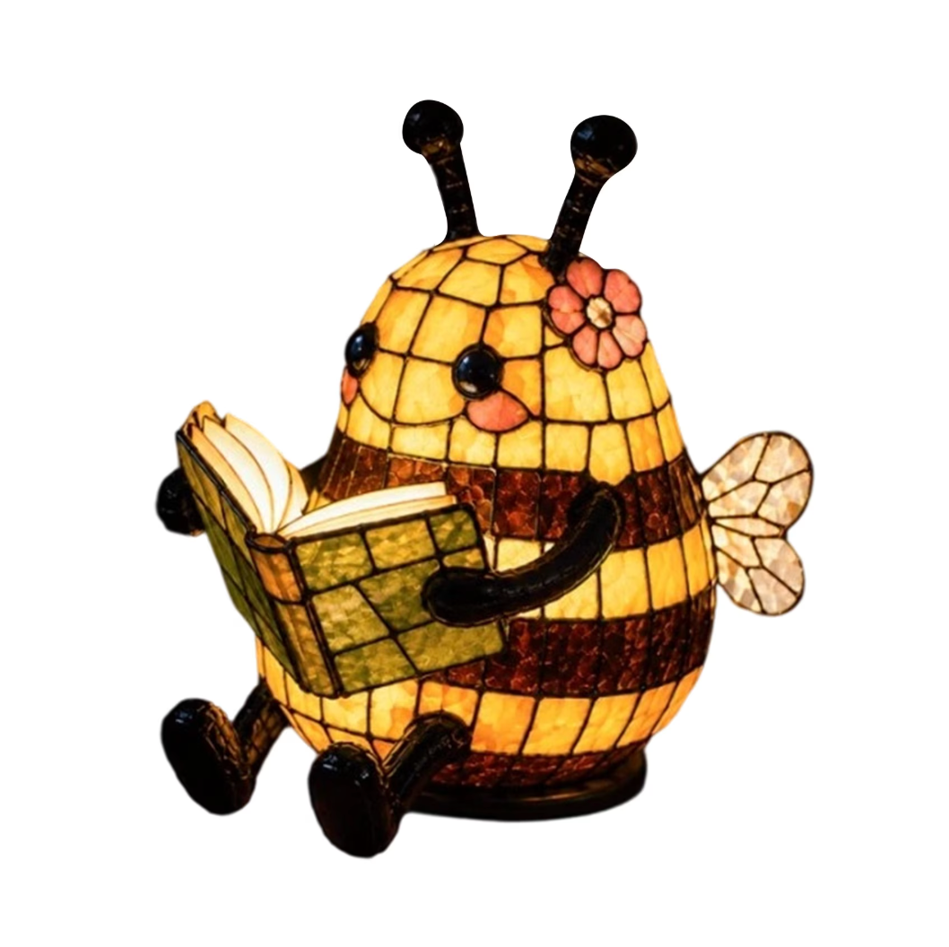 A stained-glass-style bee figurine or lamp reading an open book, with yellow and amber glass panels, dark brown outlines, black antennae and feet, a pink flower accent, and a translucent wing on a plain white background.