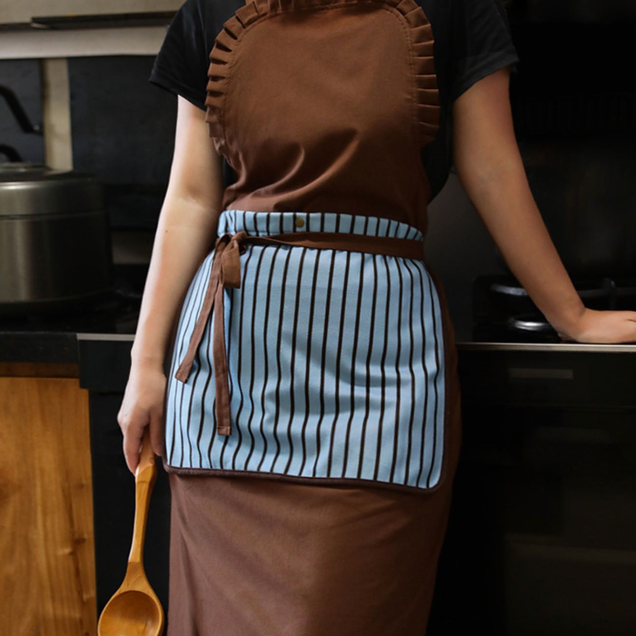 A person wearing a brown apron with a light blue and white striped front panel, standing in a kitchen and holding a wooden spoon.