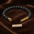 A beaded bracelet made of dark textured black beads with two gold-toned metal ends, displayed on a brown velvet surface with a small beige cylindrical piece placed below.
