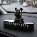 A black French bulldog figurine wearing gold sunglasses and a gold chain necklace, sitting on a black car dashboard number plate display with glowing green numbers.